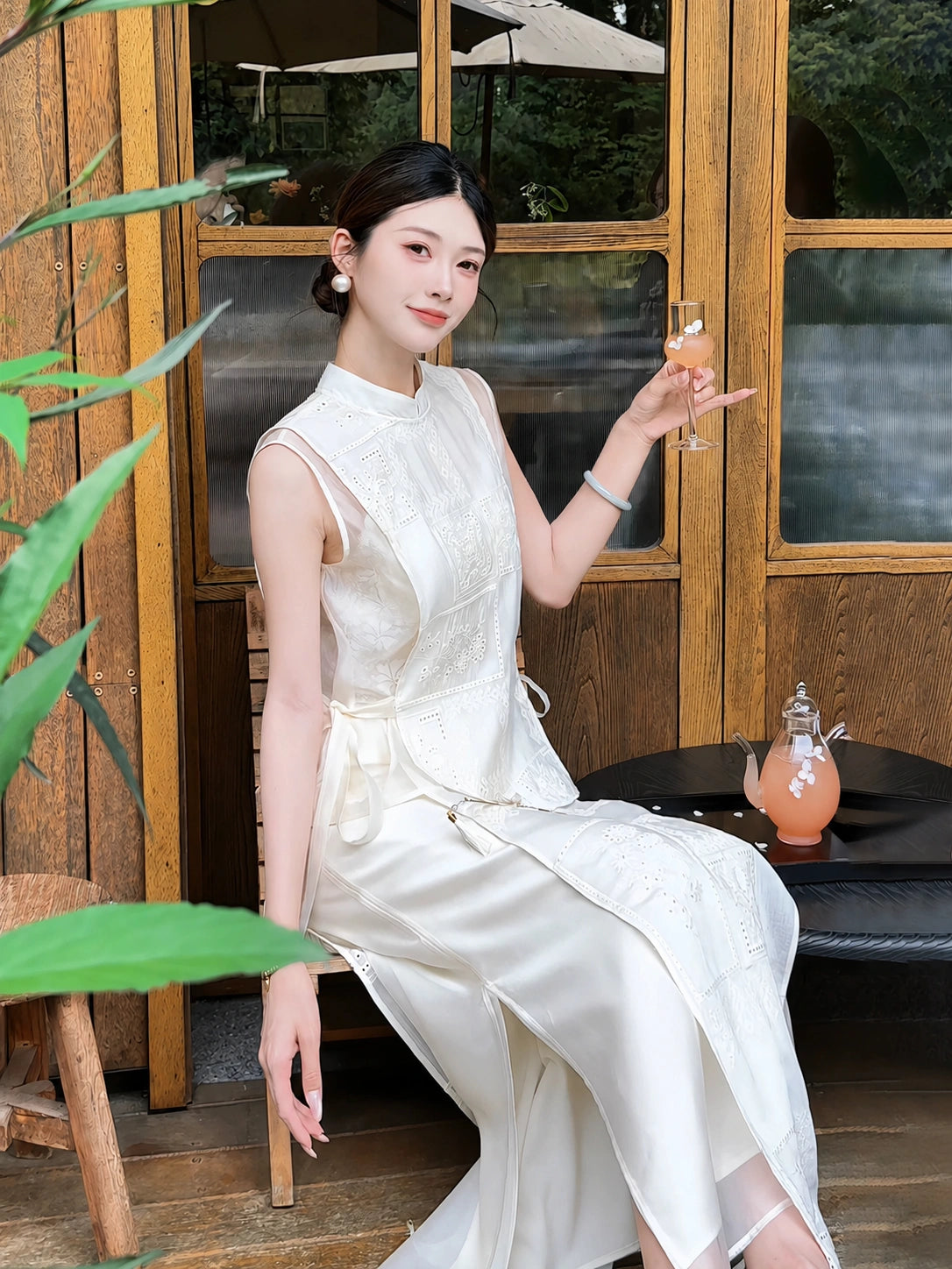 Sitting model in a cream white embroidered vest and skirt set, holding a glass of sparkling drink in a garden setting.