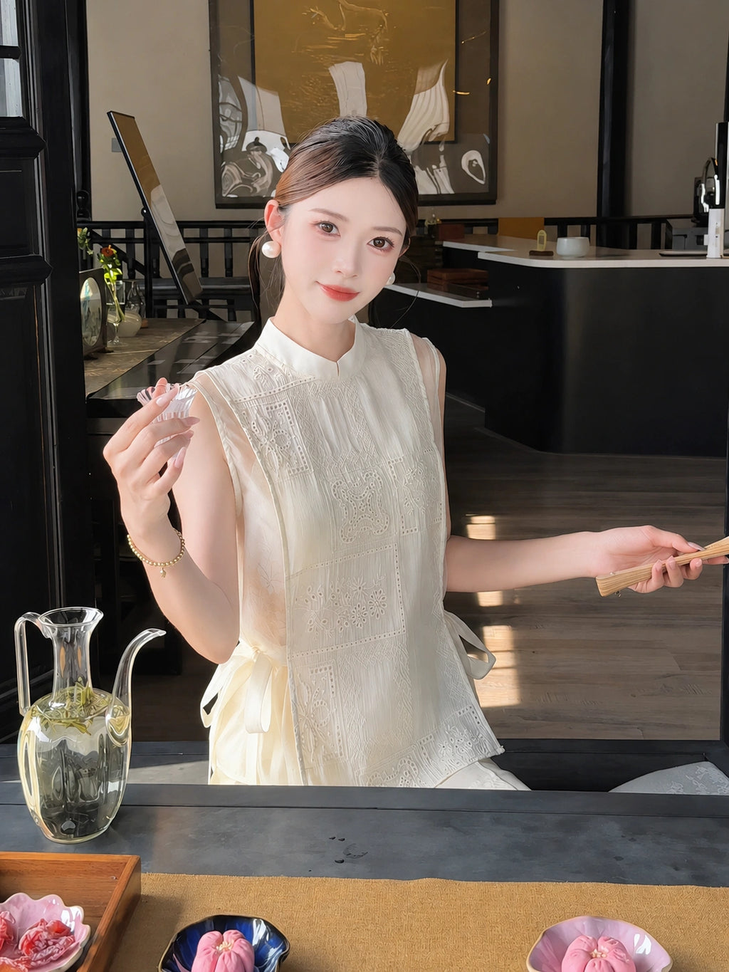 Close-up of a cream white embroidered Cheongsam vest with a mandarin collar and side ties, styled with a white skirt, holding a tea bowl.