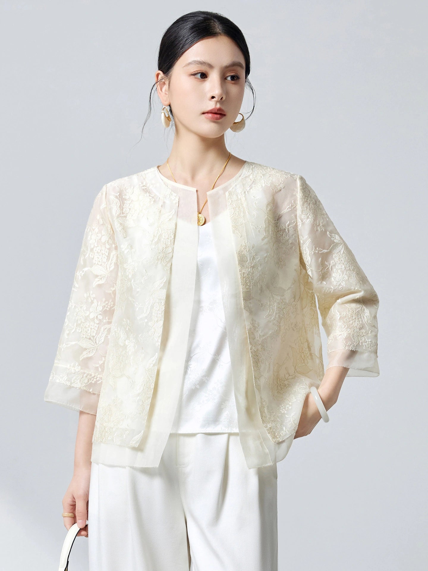 Model wearing a cream white silk organza cardigan with intricate embroidery, styled with white trousers, highlighting the versatile open-front design.