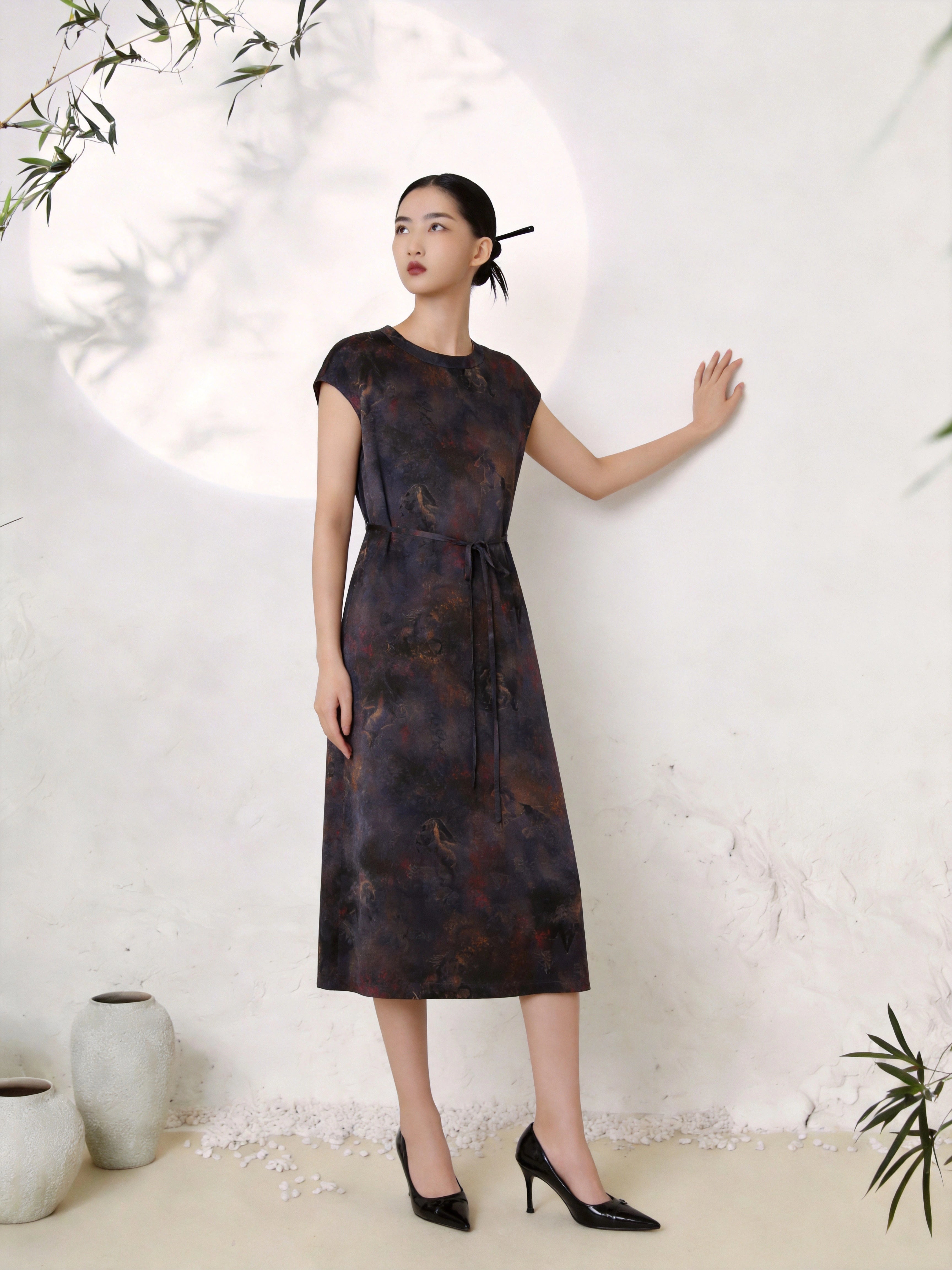 Full body shot of a dark abstract print silk midi dress with a round neckline, short cap sleeves, and a tie waist detail, crafted from 100% mulberry silk, styled with black heels.