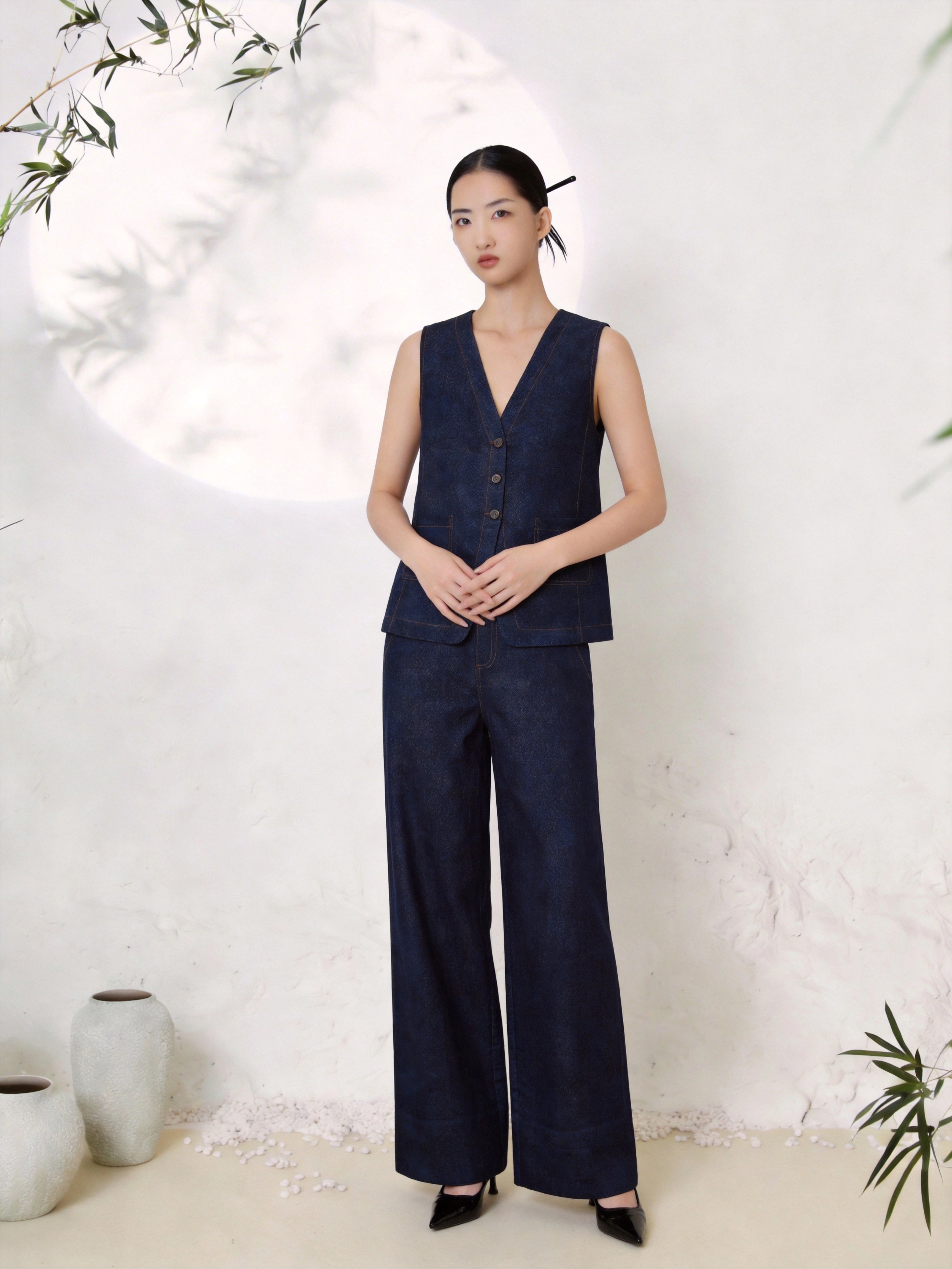 Full body shot of a deep blue silk two-piece set featuring a V-neck sleeveless vest with button-down front and contrast stitching, paired with matching wide-leg trousers, crafted from 100% mulberry silk, styled with black heels.