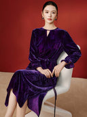 Seated model wearing the purple velvet dress, showing its draped skirt, floral waist tie and ruffled cuffs.