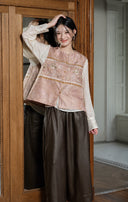 Model posing beside a mirror in the rose vest, cream blouse and brown trousers.