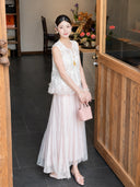 Model wearing a white embroidered vest with a ruffled hem, paired with a pink pleated maxi skirt, holding a pink handbag, 100% polyester.