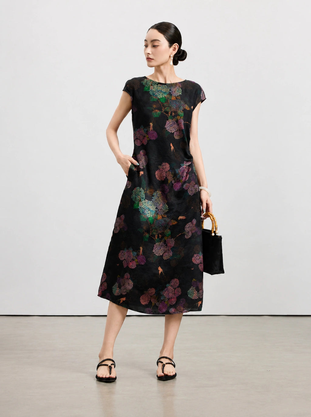 Black floral silk midi dress styled with a bamboo-handle handbag and flat sandals.