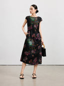 Black floral silk midi dress styled with a bamboo-handle handbag and flat sandals.