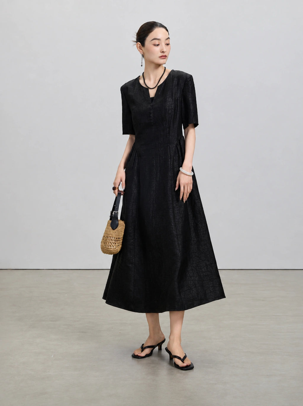 Model wearing the black fit-and-flare Xiangyunsha midi dress with a woven handbag