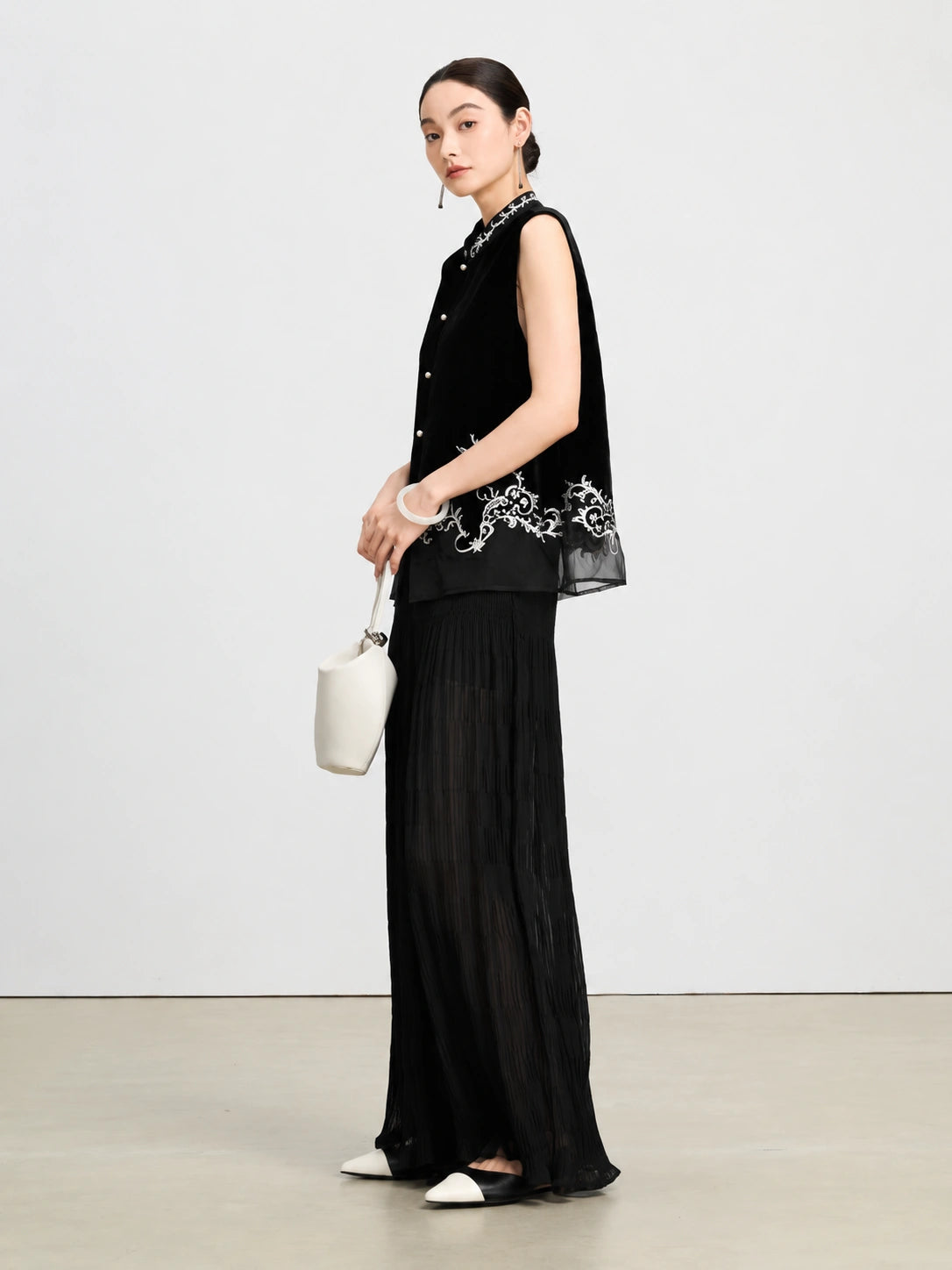 Model wearing a black embroidered silk velvet vest with a sheer hem and wide-leg trousers.