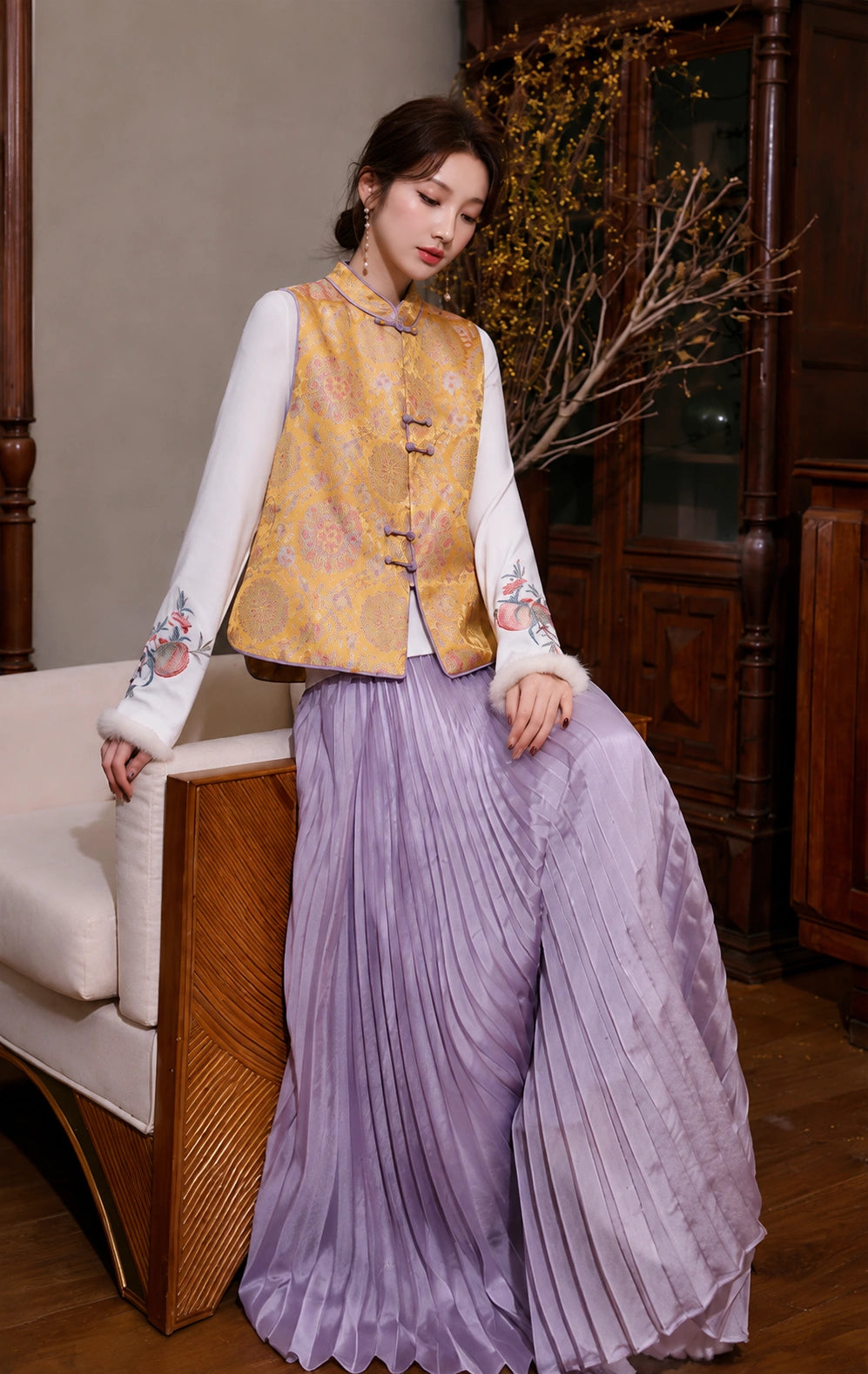 Seated model styling the golden New Chinese brocade vest over an embroidered white top with lavender pleated trousers.