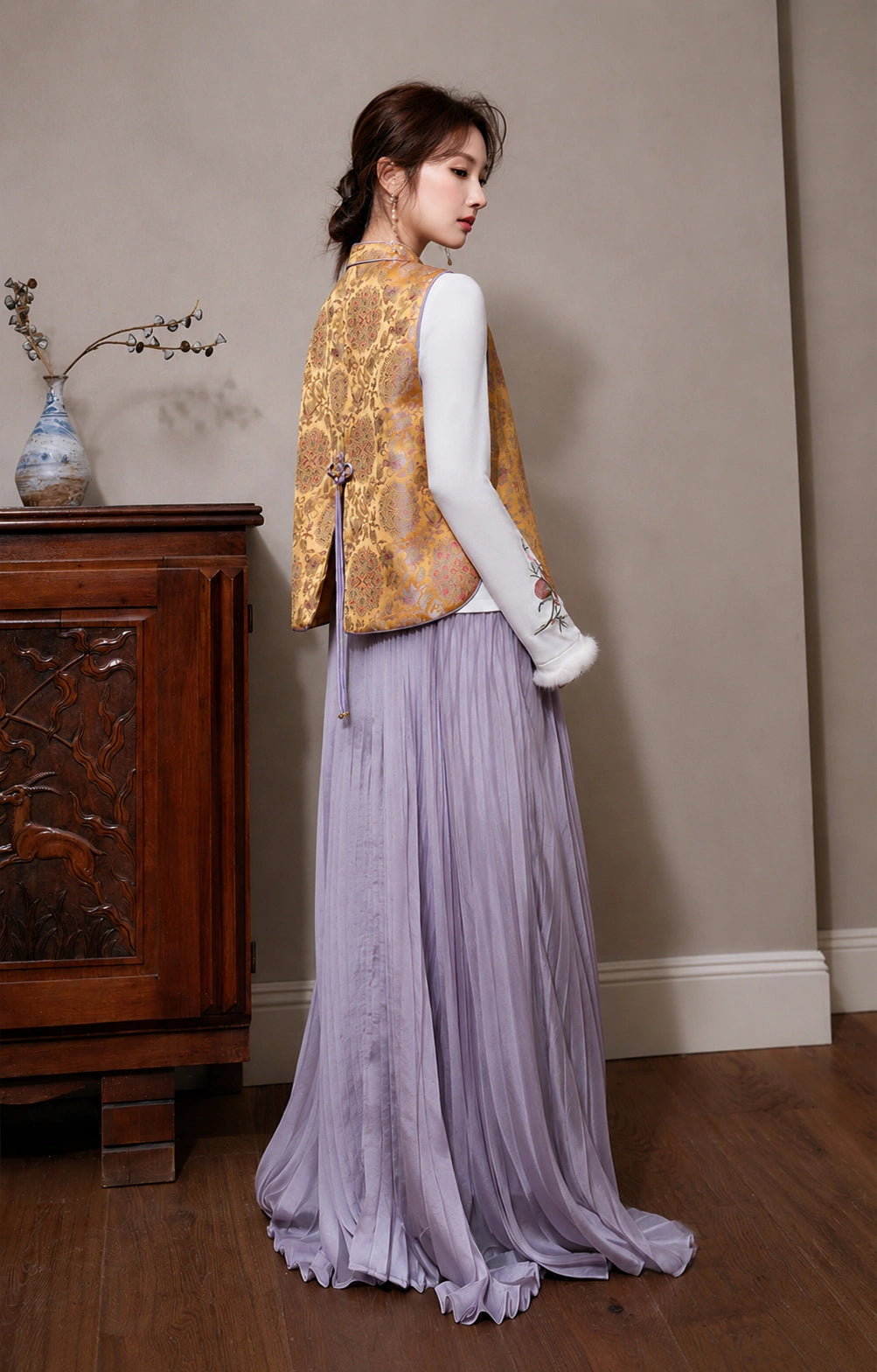 Model wearing the golden floral layering vest from the back with a white long-sleeve top and lavender pleated skirt.