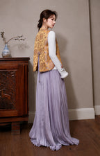 Model wearing the golden floral layering vest from the back with a white long-sleeve top and lavender pleated skirt.