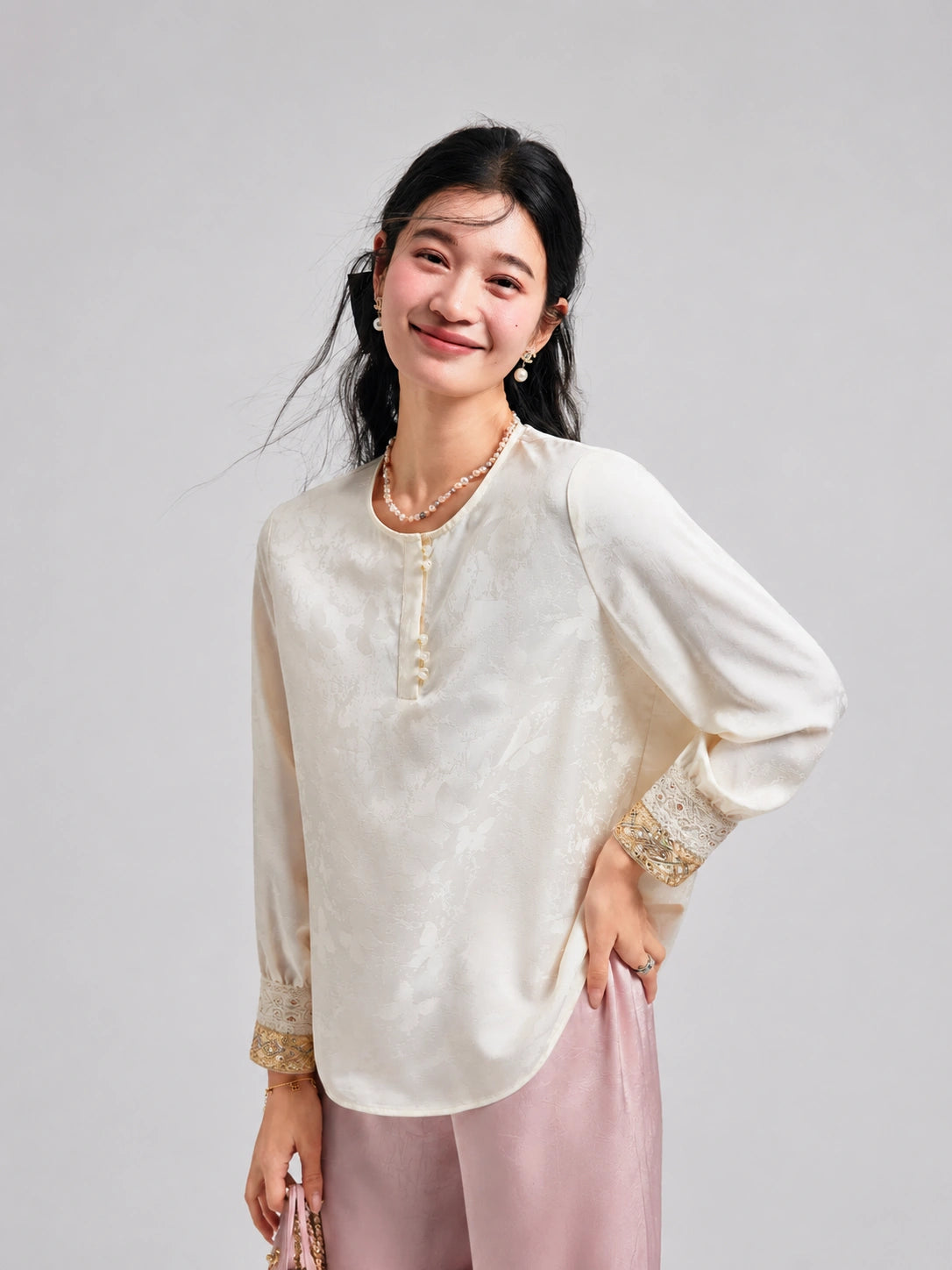 Model wearing an ivory long-sleeve satin top with a round neckline and gold-accented beaded cuffs.