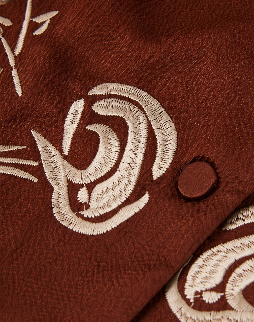 Close-up of ivory floral embroidery and a silk-covered button on rust-brown gambiered silk.