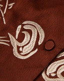 Close-up of ivory floral embroidery and a silk-covered button on rust-brown gambiered silk.