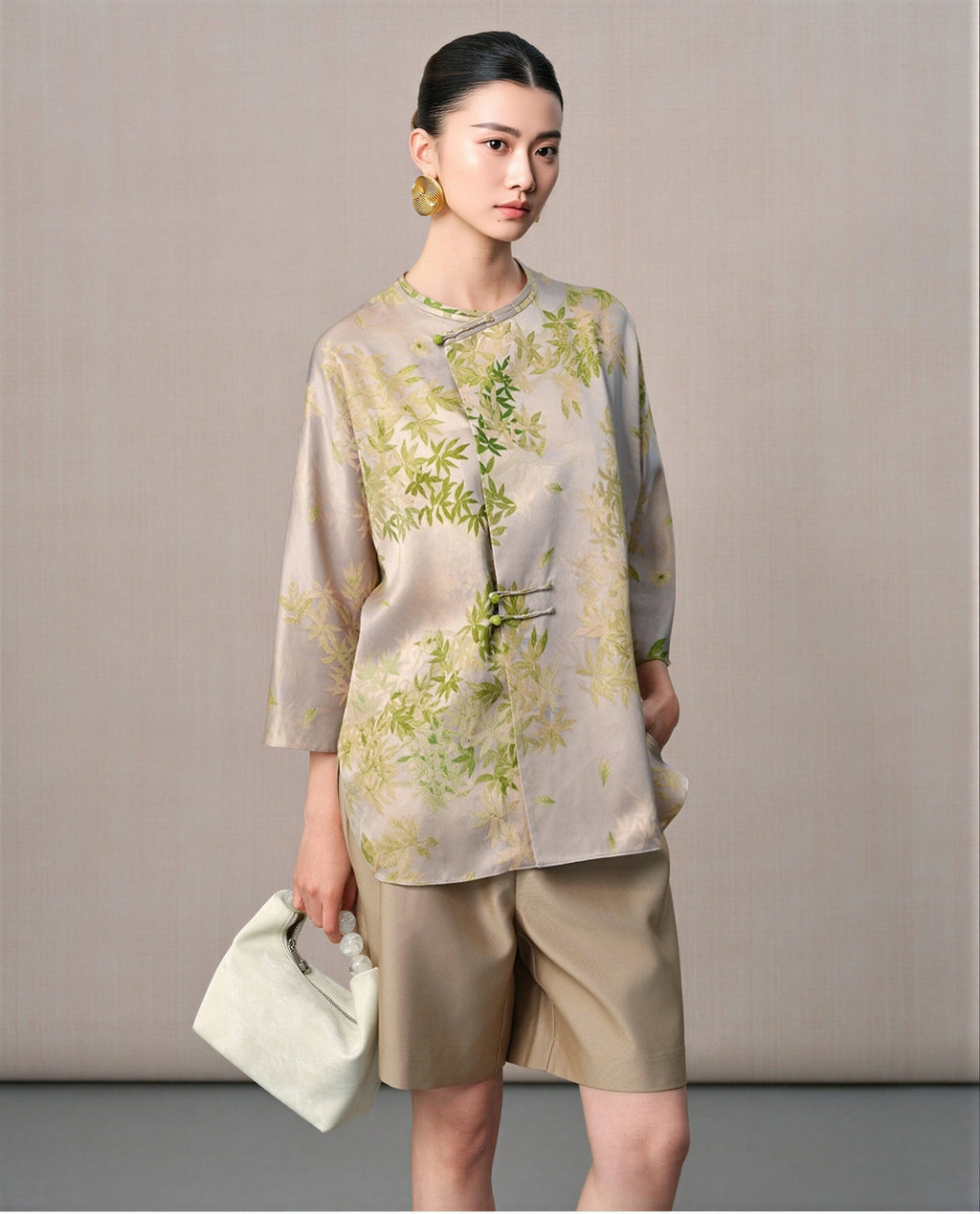 Model wearing a taupe and sage green leaf-print silk blouse with a round neckline and asymmetric button detail
