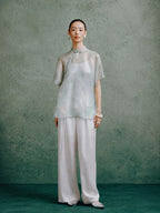 Women's light celadon silk cheongsam top paired with wide-leg white trousers, styled in a modern Chinese-inspired outfit.