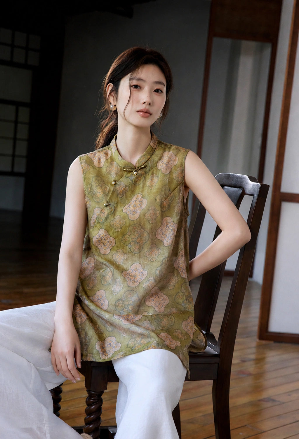 Seated model wearing an olive jacquard long vest with Mandarin collar and decorative closures