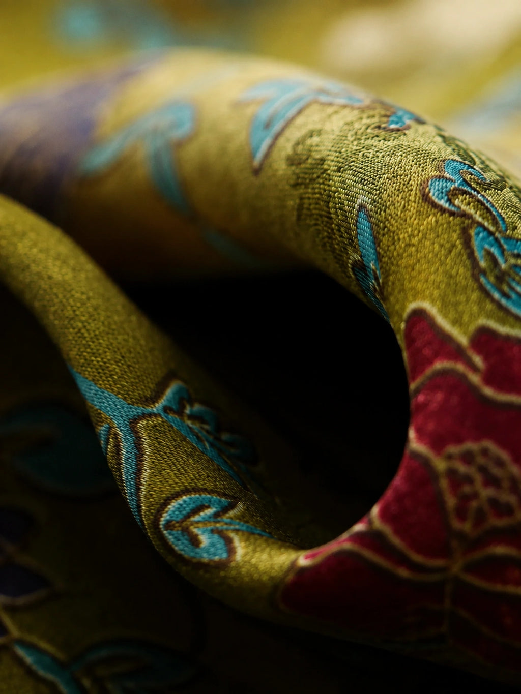 Close-up of lustrous olive-gold fabric with turquoise botanical motifs and deep red floral accents
