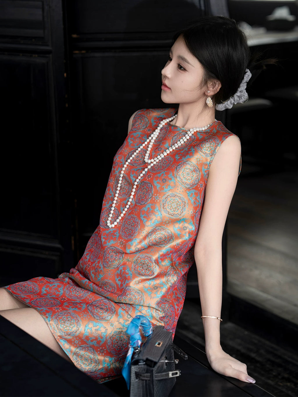Seated model wearing a lightweight vermilion and turquoise Shu brocade sleeveless dress.
