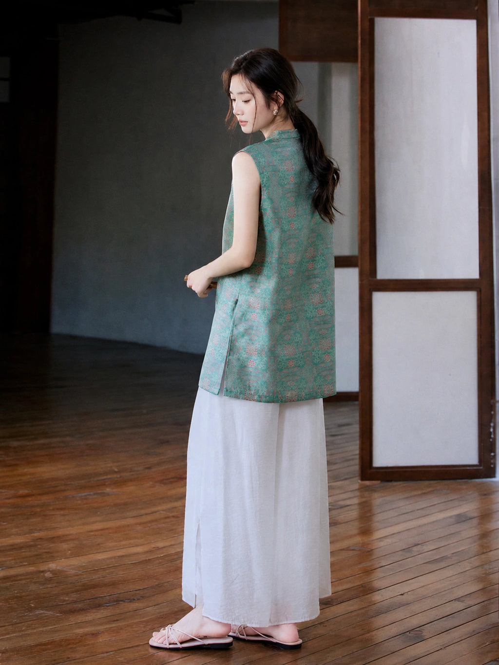 Back three-quarter view of the teal and pink Song brocade longline vest styled with white trousers