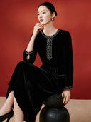 Seated model wearing the black velvet midi dress with embroidered neckline, cuffs and tie waist