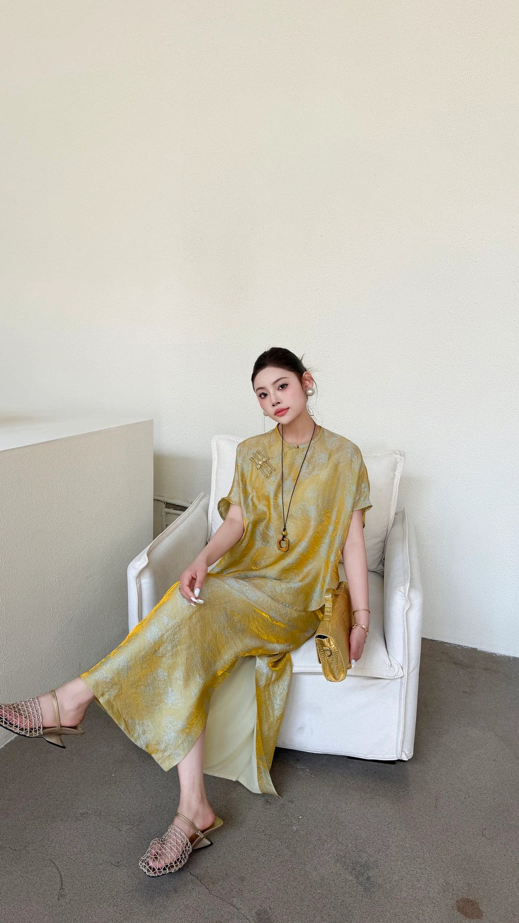 Woman sitting in a white armchair wearing a gold Qipao top and matching long skirt with side slits, showcasing the relaxed fit and soft drape of the viscose blend fabric.