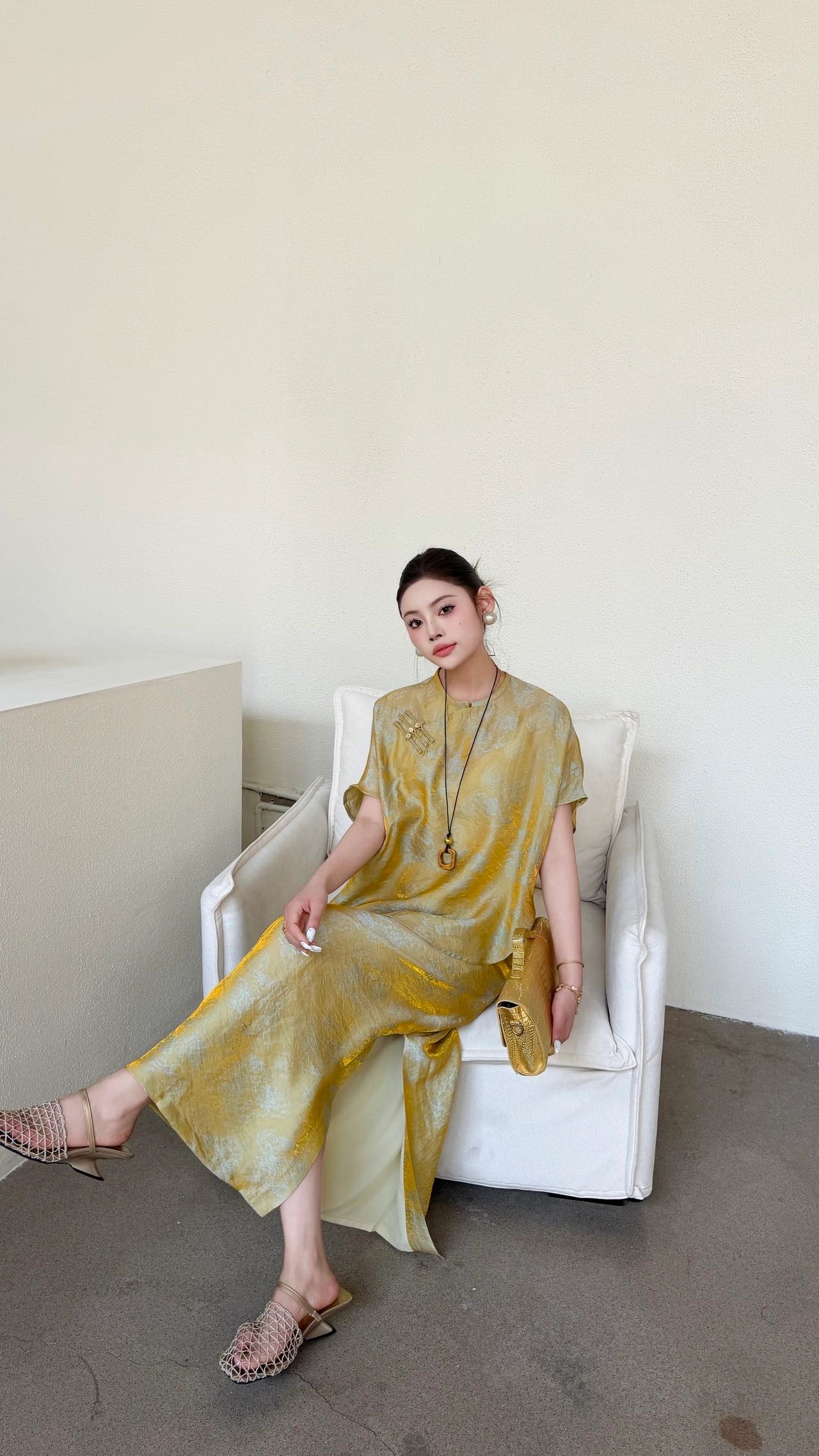 Woman sitting in a white armchair wearing a gold Qipao top and matching long skirt with side slits, showcasing the relaxed fit and soft drape of the viscose blend fabric.