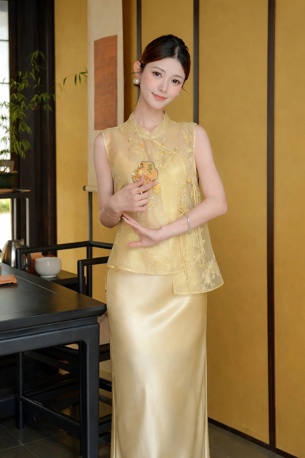 Model wearing a gold sleeveless embroidered vest with a mandarin collar and side ties, paired with a matching satin skirt, holding a fan in a traditional setting.