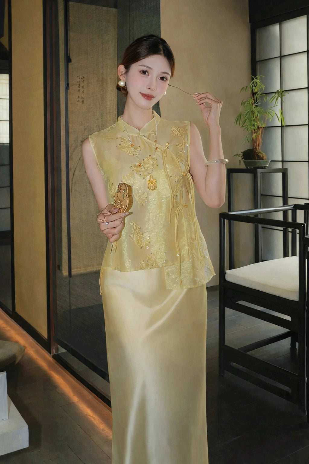 Model in a gold sleeveless embroidered vest with a mandarin collar, holding a fan and touching her hair, showcasing the intricate embroidery.