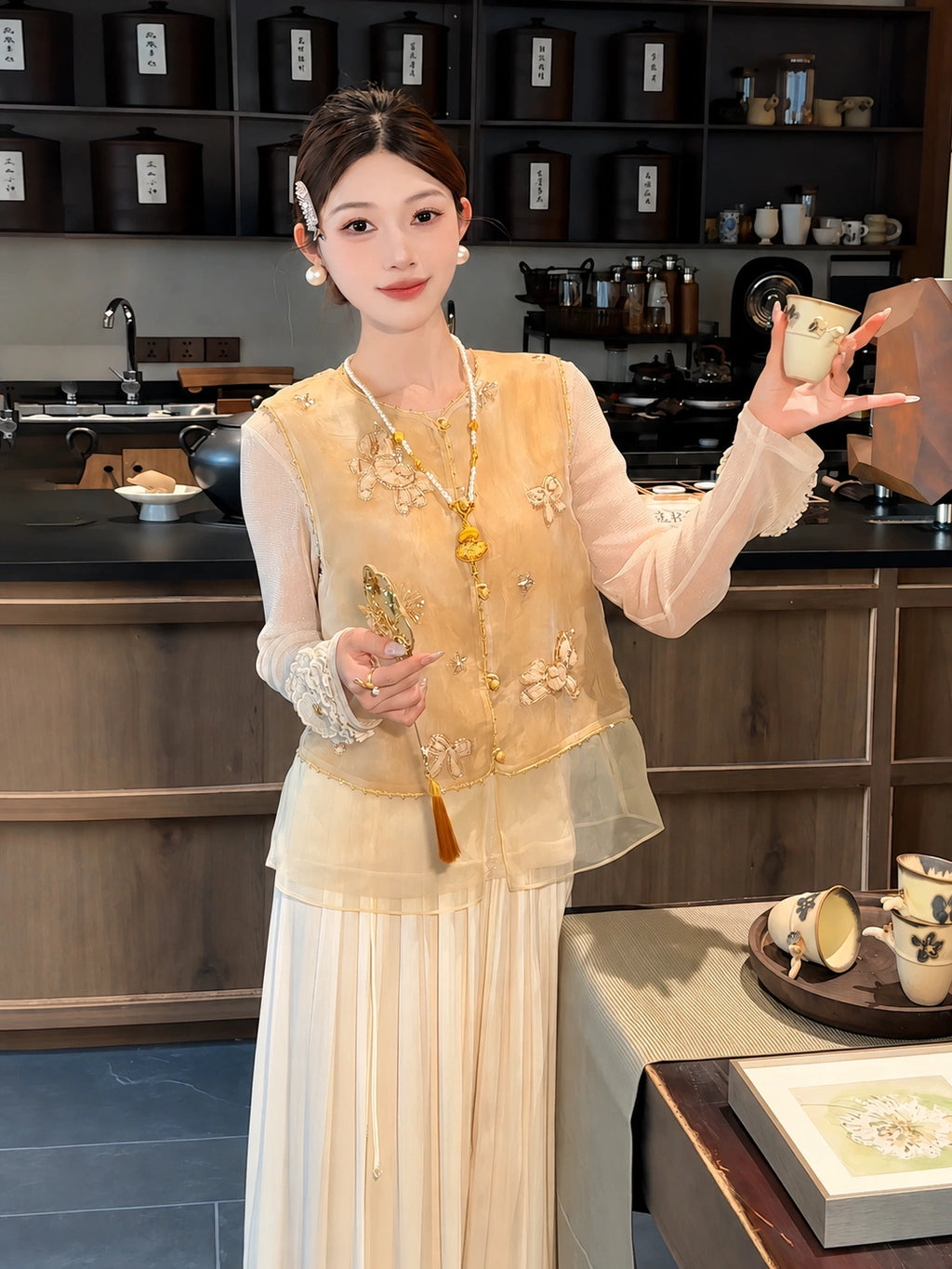 Model wearing a gold sleeveless embroidered vest with beaded details, styled over a sheer top and white pleated skirt, holding a fan and tea cup.