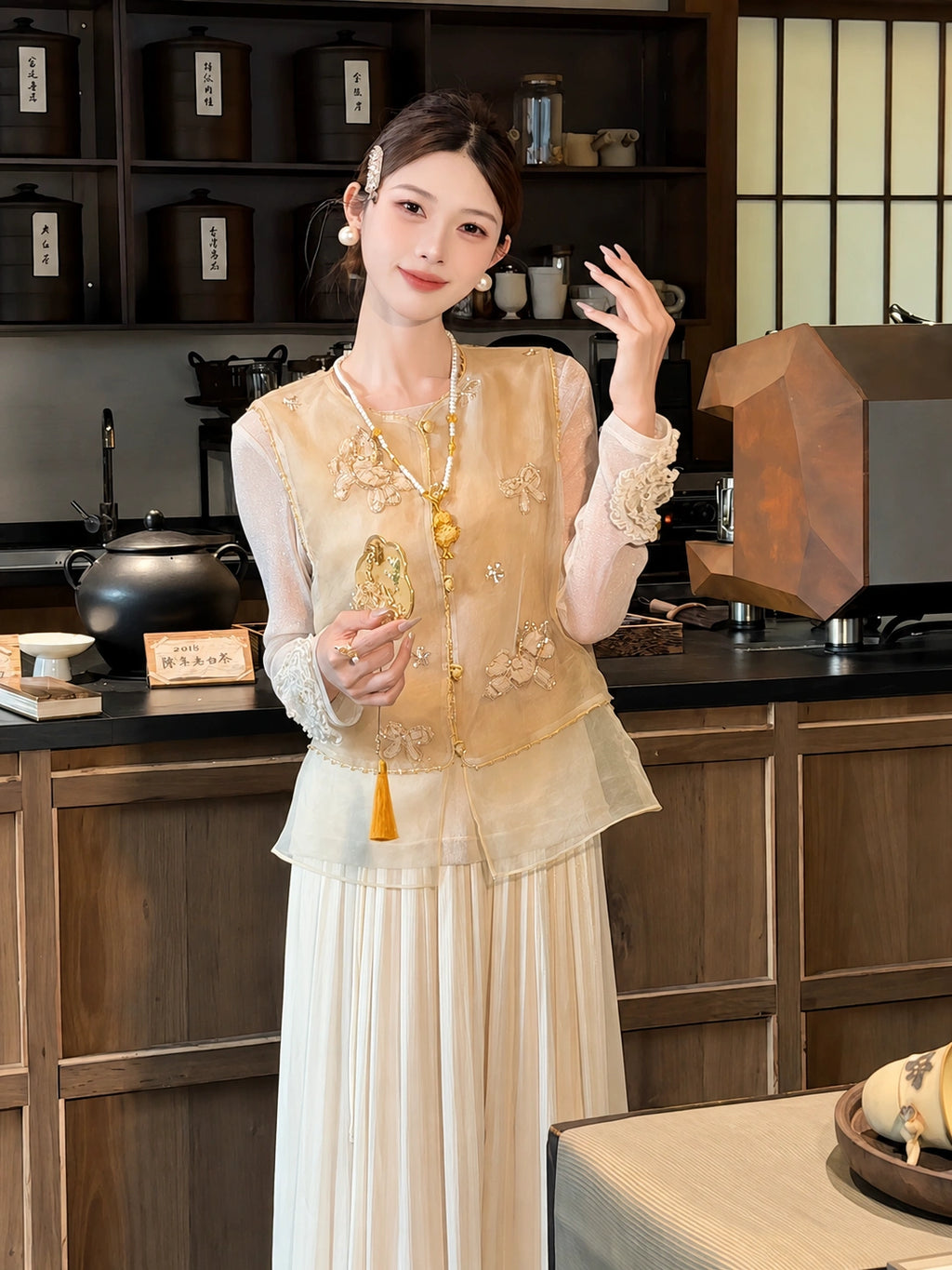 Front view of a gold embroidered vest with beadwork and a layered sheer hem, paired with a white pleated skirt and a long pearl necklace.