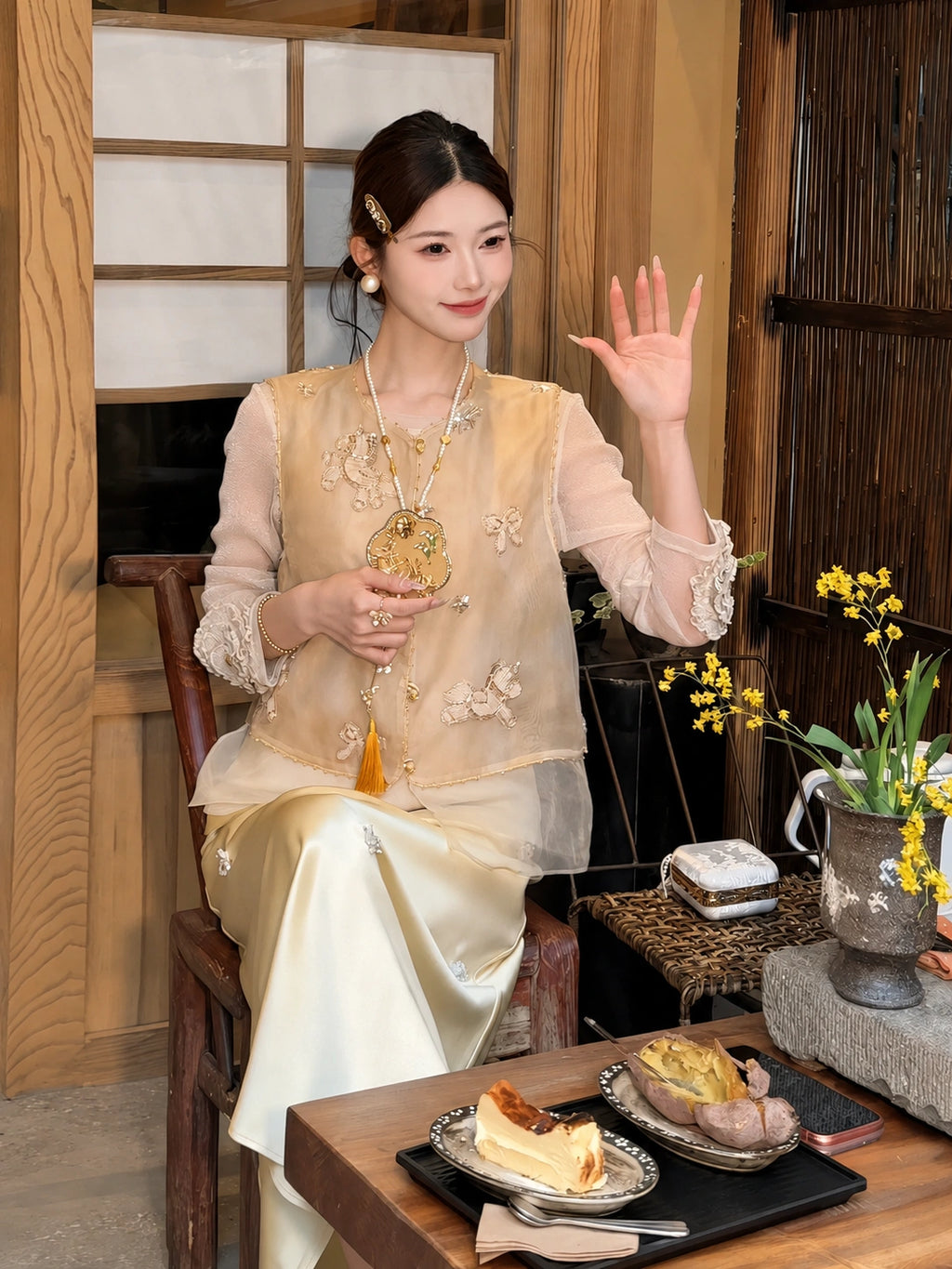 Sitting model in a gold beaded vest with a layered sheer hem, styled with a cream skirt and holding a gold fan, showcasing the relaxed fit and elegant details.