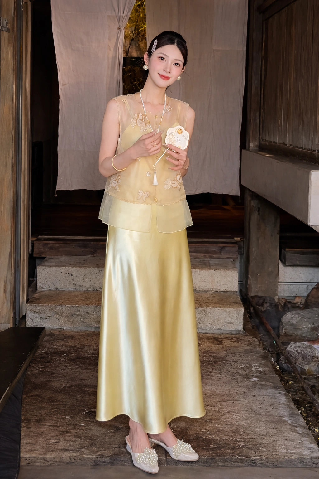 Model in a gold sheer embroidered top with a tassel necklace, paired with a matching satin maxi skirt, holding a traditional fan.