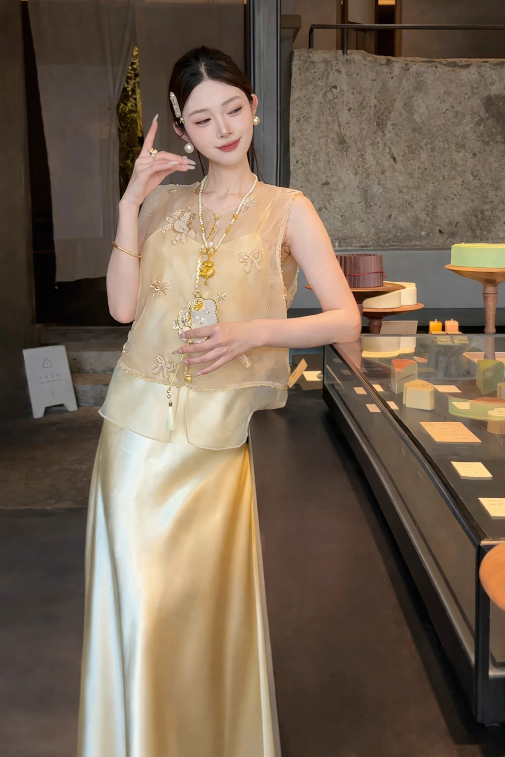 Side profile of a woman wearing a gold floral embroidered mesh top and satin skirt, standing by a glass display counter.