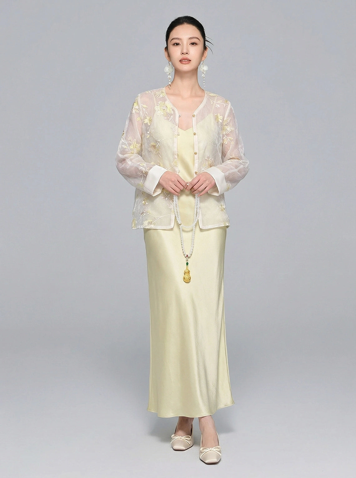 Model wearing a sheer V-neck New Chinese style shirt with intricate yellow/gold floral embroidery and sequins, paired with a matching satin skirt, holding a long pearl necklace.