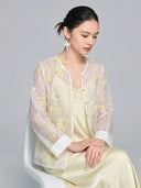 Seated model in a sheer embroidered shirt with gold floral details and sequins, featuring white cuffs and front buttons, styled with a satin slip dress.