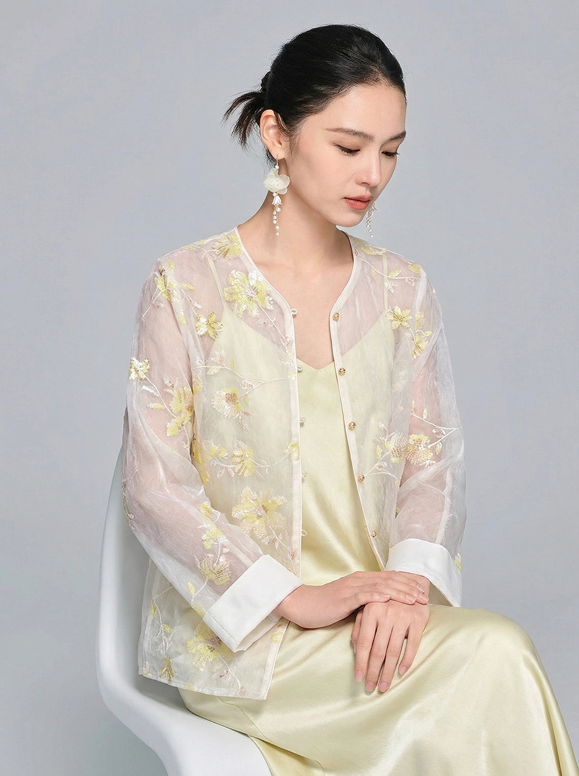 Seated model in a sheer embroidered shirt with gold floral details and sequins, featuring white cuffs and front buttons, styled with a satin slip dress.