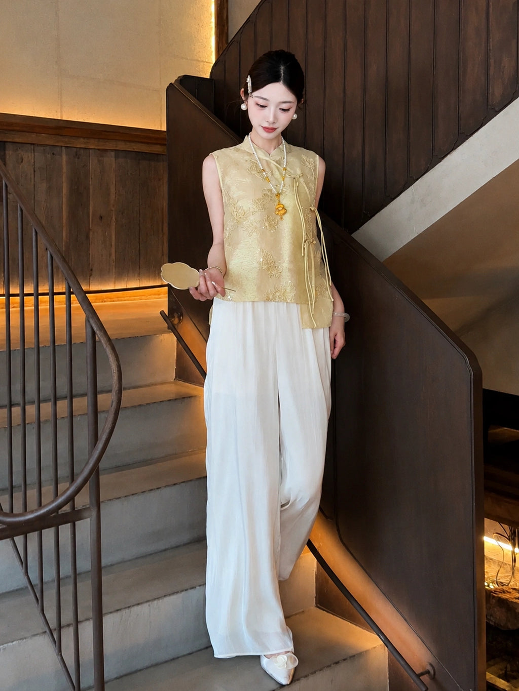 Model wearing a gold embroidered Cheongsam vest paired with white pleated wide-leg pants, holding a fan on stairs.