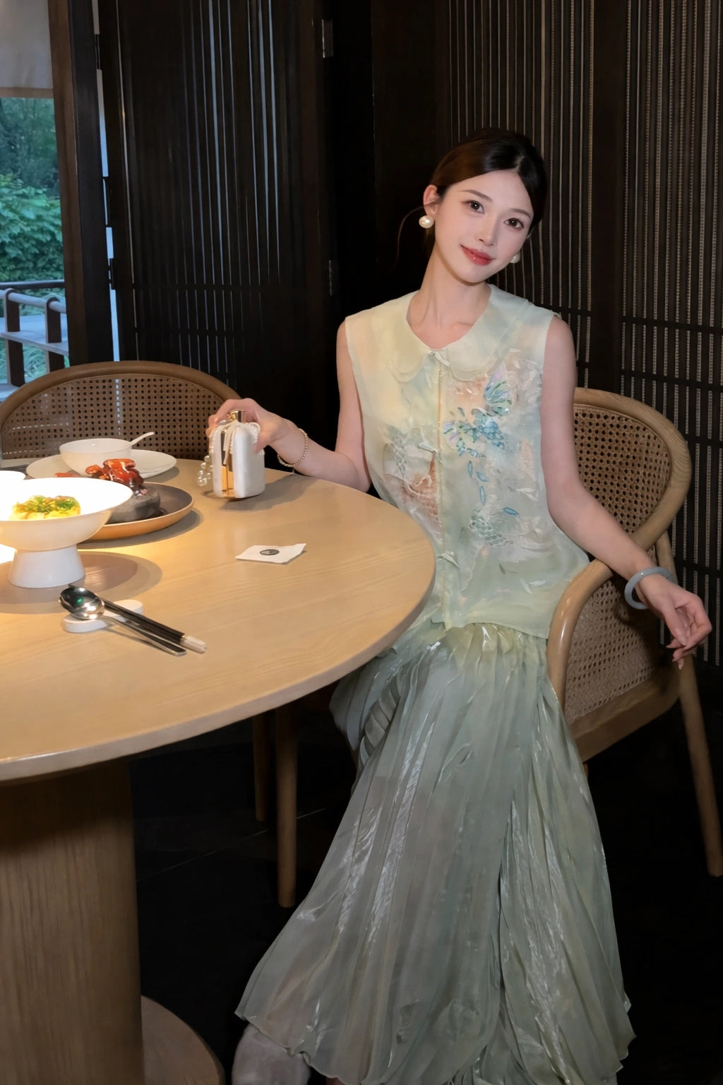 Sitting model in a green silk-blend vest with floral embroidery and pearl buttons, holding a small object, showcasing the relaxed fit and elegant drape.