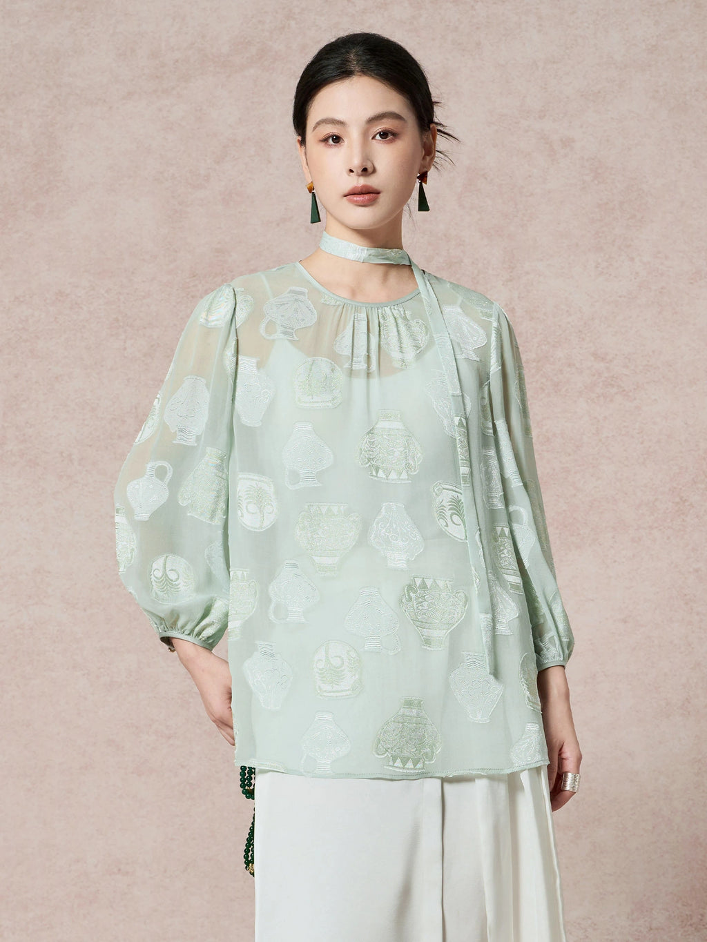 Model wearing a mint green 100% mulberry silk blouse with a round neck, sheer cutwork pattern, and a matching scarf tie, styled with white pleated pants.