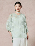 Model wearing a mint green 100% mulberry silk blouse with a round neck, sheer cutwork pattern, and a matching scarf tie, styled with white pleated pants.