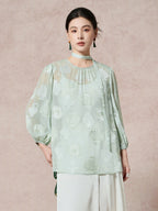 Model wearing a mint green 100% mulberry silk blouse with a round neck, sheer cutwork pattern, and a matching scarf tie, styled with white pleated pants.