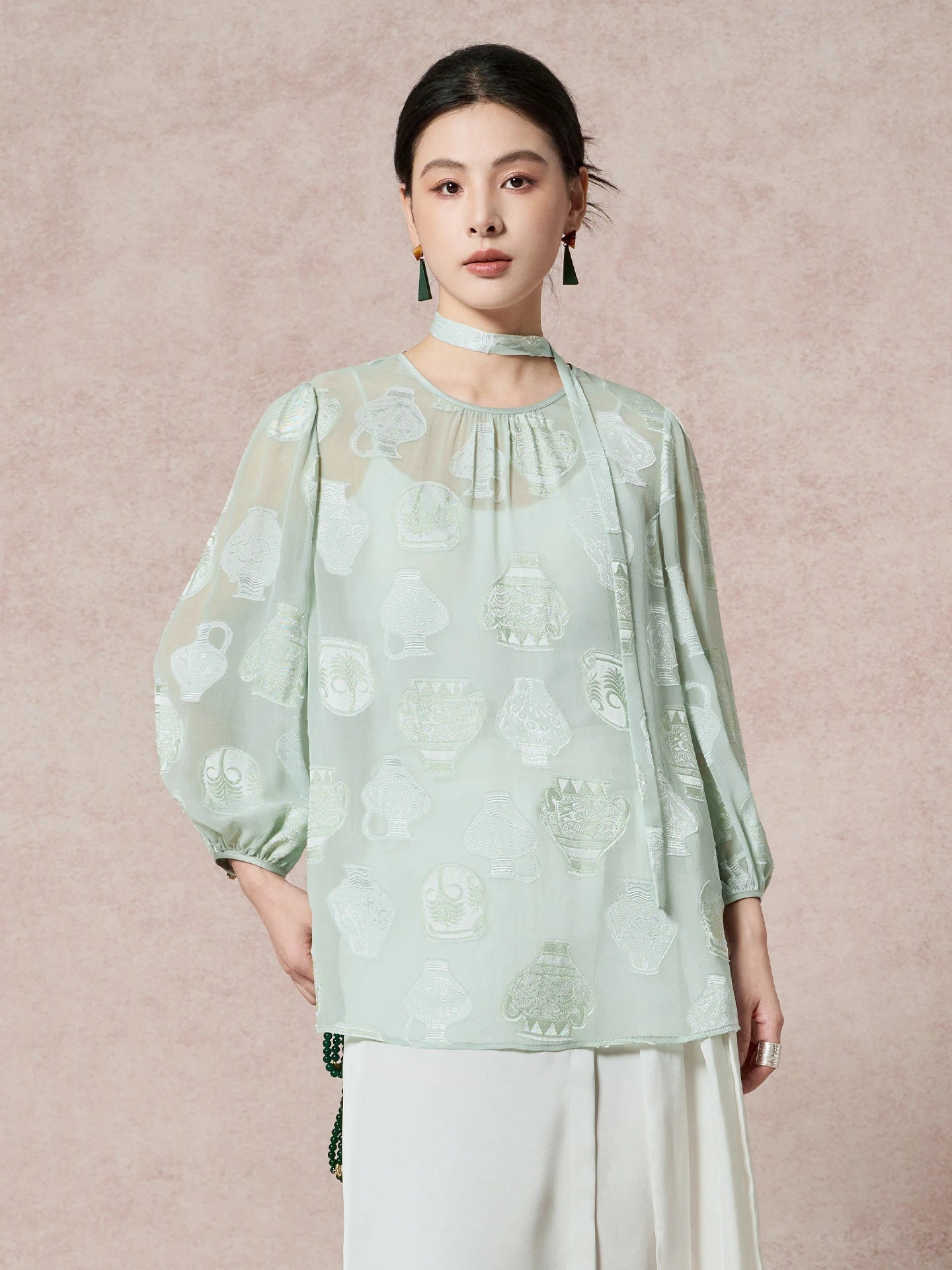 Model wearing a mint green 100% mulberry silk blouse with a round neck, sheer cutwork pattern, and a matching scarf tie, styled with white pleated pants.