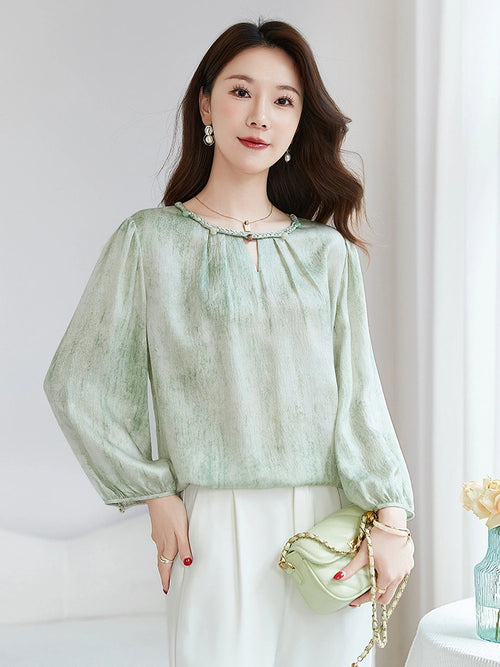 Side view of a model wearing a green silk blouse with a braided neckline, sitting on a chair, showcasing the relaxed fit and flowy drape.