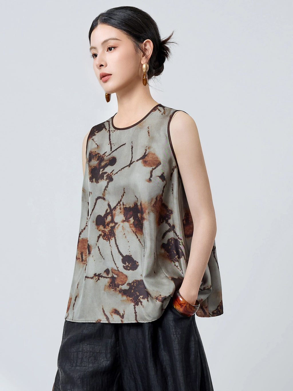 Side profile of a women's grey silk tank top with brown piping and an abstract floral pattern, styled with black pants and a brown bag.