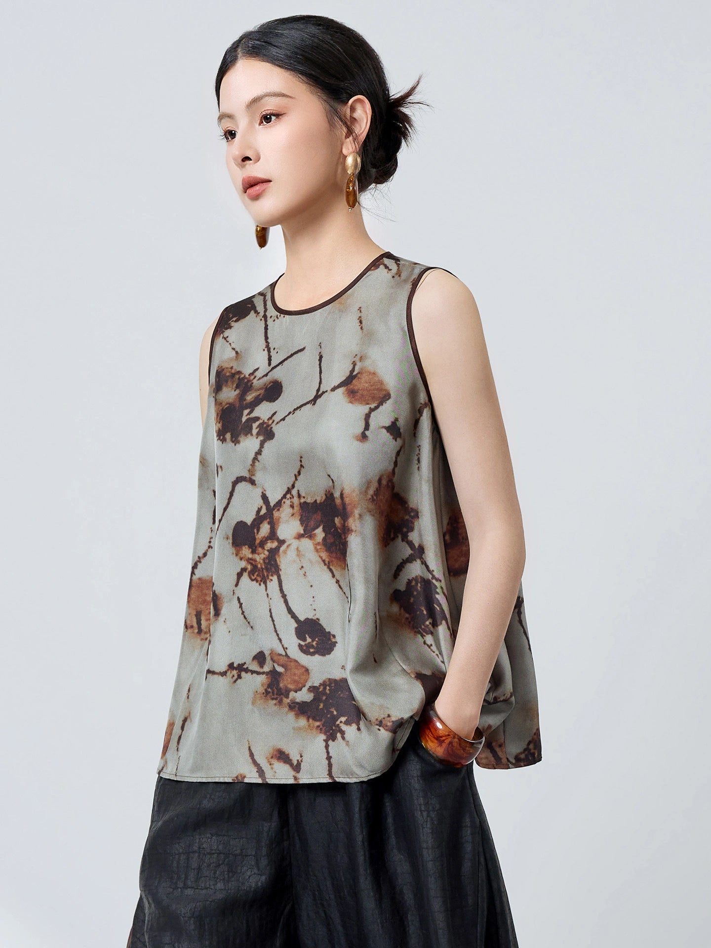 Side profile of a women's grey silk tank top with brown piping and an abstract floral pattern, styled with black pants and a brown bag.