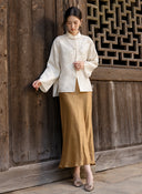 Full-length outfit featuring an ivory embellished jacket, a flowing bronze skirt and metallic shoes beside a wooden lattice window.