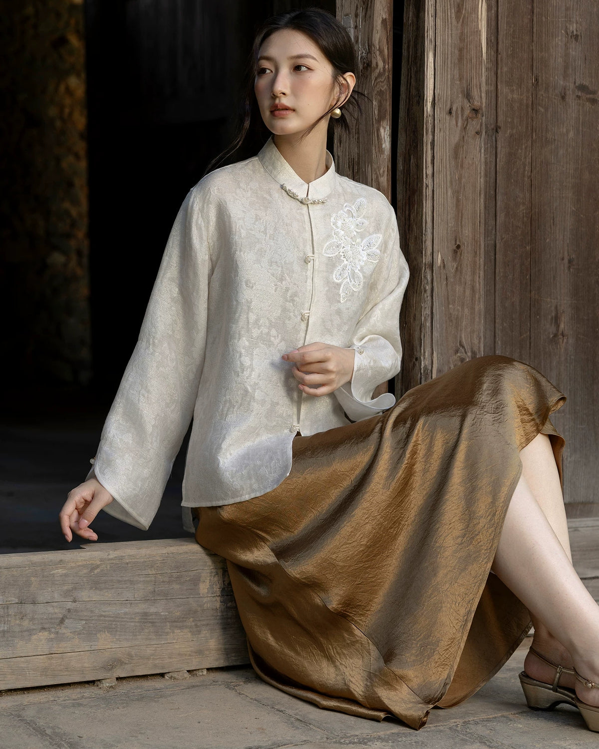 Model seated beside a wooden wall wearing an ivory embellished jacquard jacket with a bronze skirt and metallic shoes.