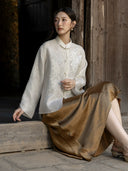 Model seated beside a wooden wall wearing an ivory embellished jacquard jacket with a bronze skirt and metallic shoes.