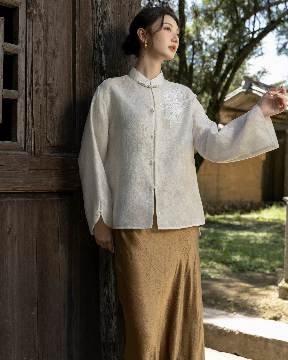 Model raising one arm to show the wide sleeve and split cuff of an ivory jacket with floral chest embellishment.